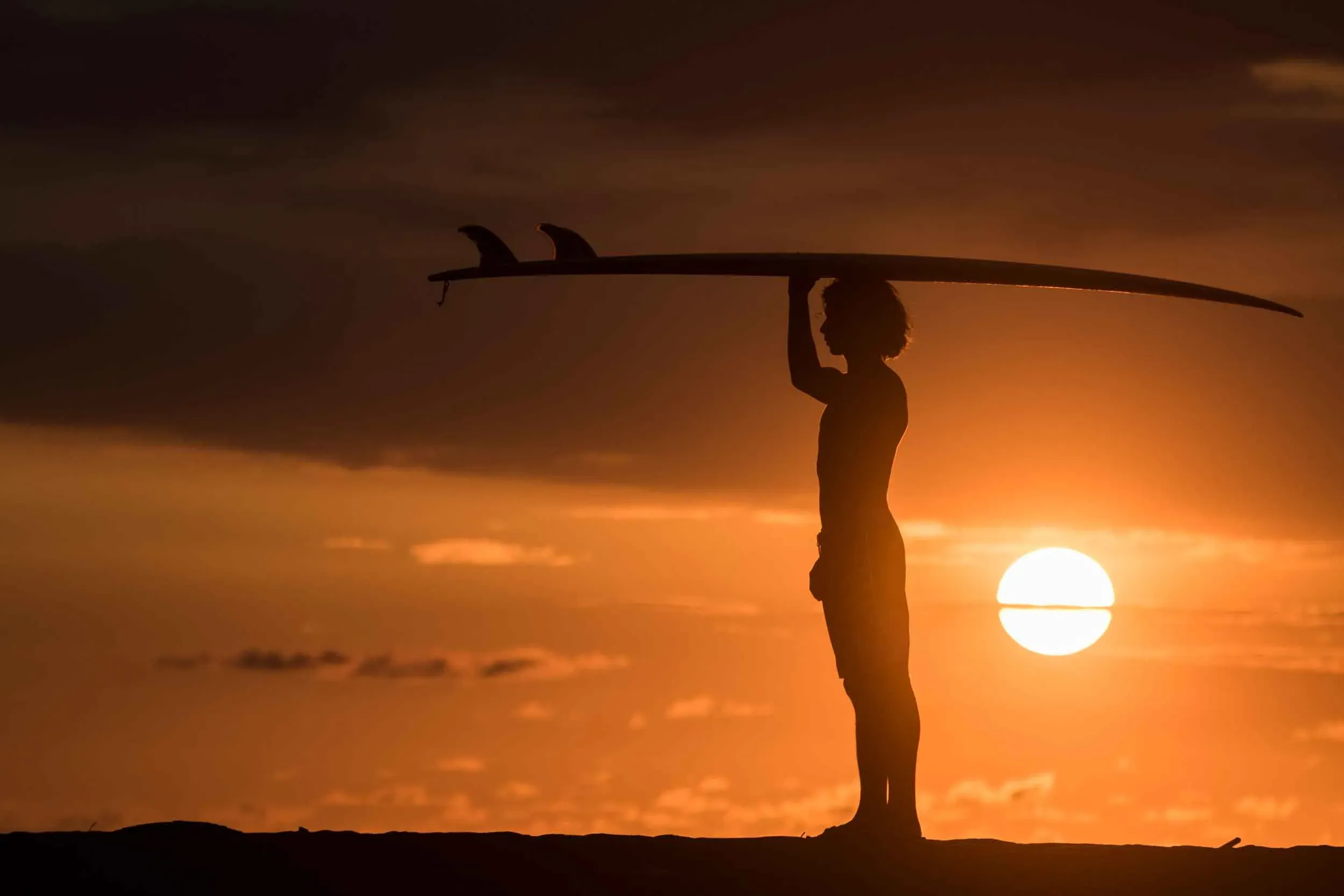 "Beautiful sunset with a young person holding a surfboard on their head, gazing at the colorful sky."