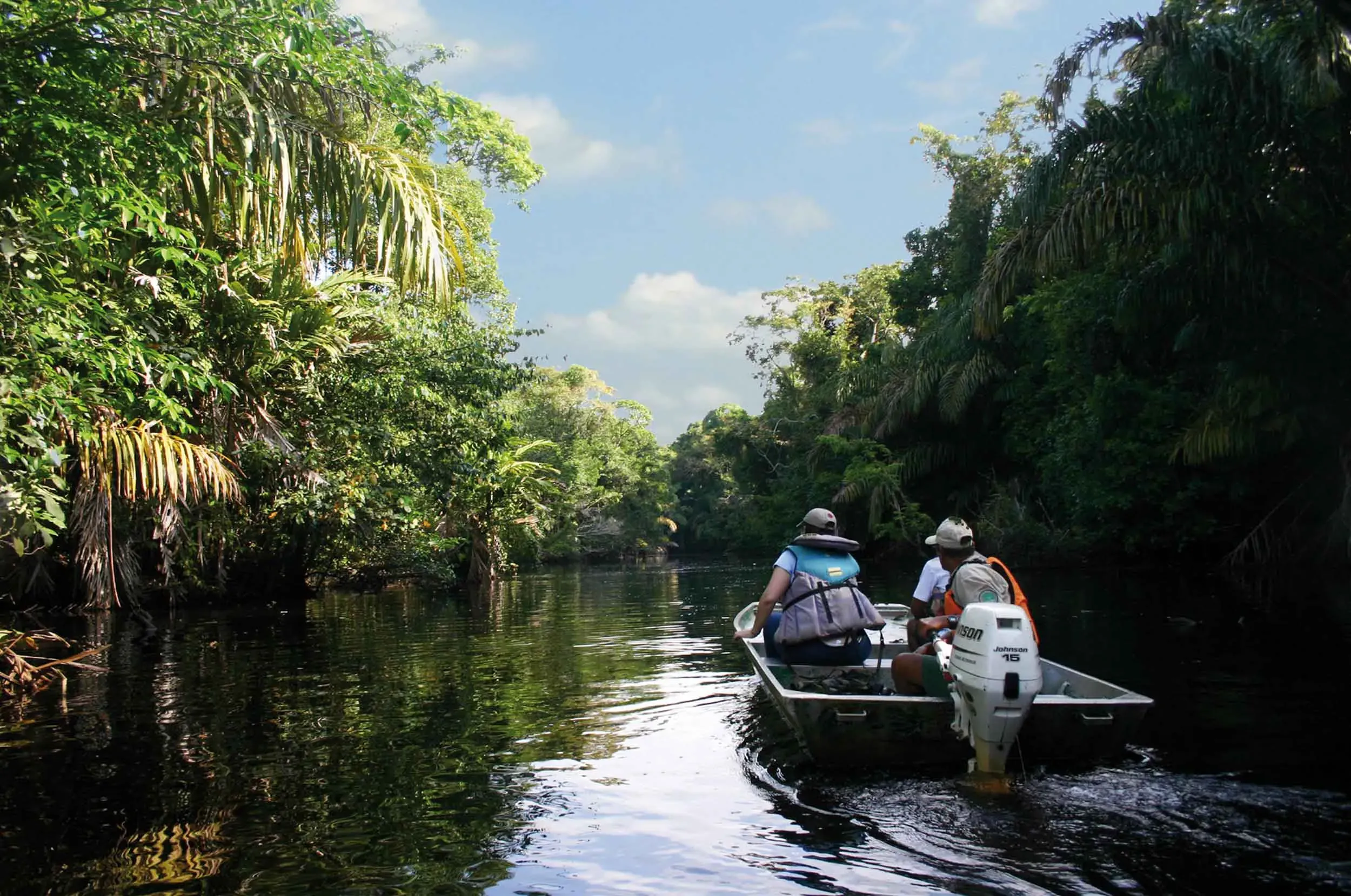 Boat with three people in Tortuguero National Park on a sunny day, surrounded by lush forests.